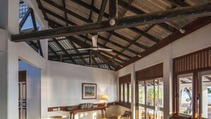 A spacious room with a high ceiling, wooden beams, a sofa, chairs, a small table, and large windows allowing natural light. - Moon Isle Beach Bunglow (Nilaveli)