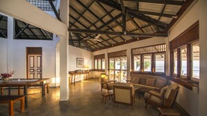 A spacious room with a high ceiling, wooden furniture, and large windows offering views of the outdoors. - Moon Isle Beach Bunglow (Nilaveli)