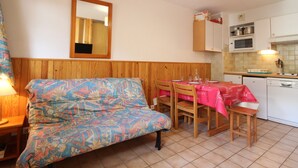 A small, cozy room with a wooden headboard, a sofa with a colorful patterned cover, a dining table with chairs, and a kitchen area with cabinets and a microwave.