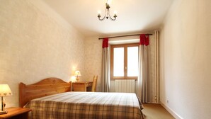 A bedroom with a wooden bed, a nightstand with a lamp, a chair, a window with curtains, and a radiator. - Spacious apartment 5 ppl with balcony and wifi in Lanslebourg (Lanslebourg-Mont-Cenis)