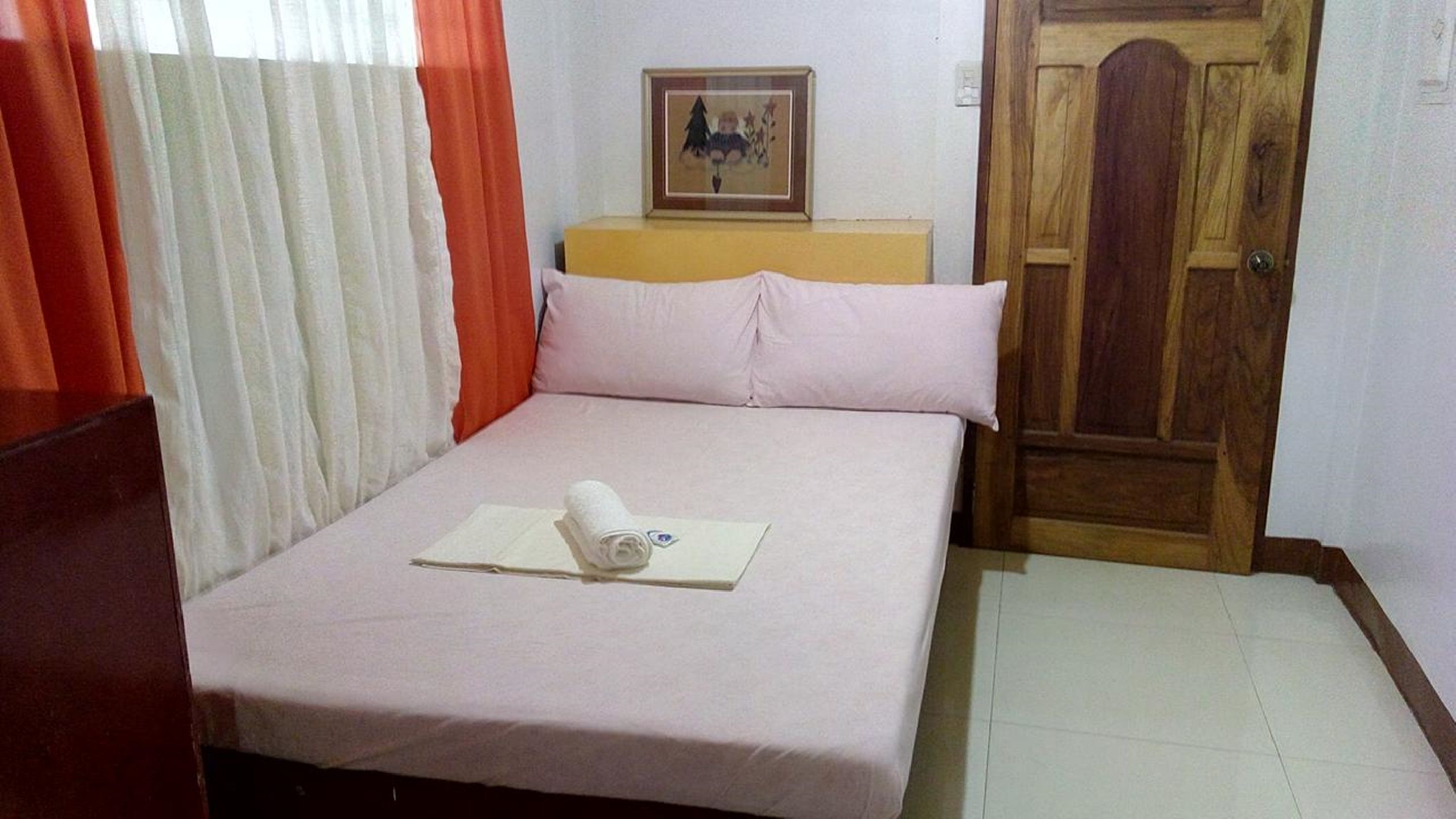 A small, neatly made bed with a white sheet and a rolled towel, a wooden nightstand with a framed picture, and a wooden door.