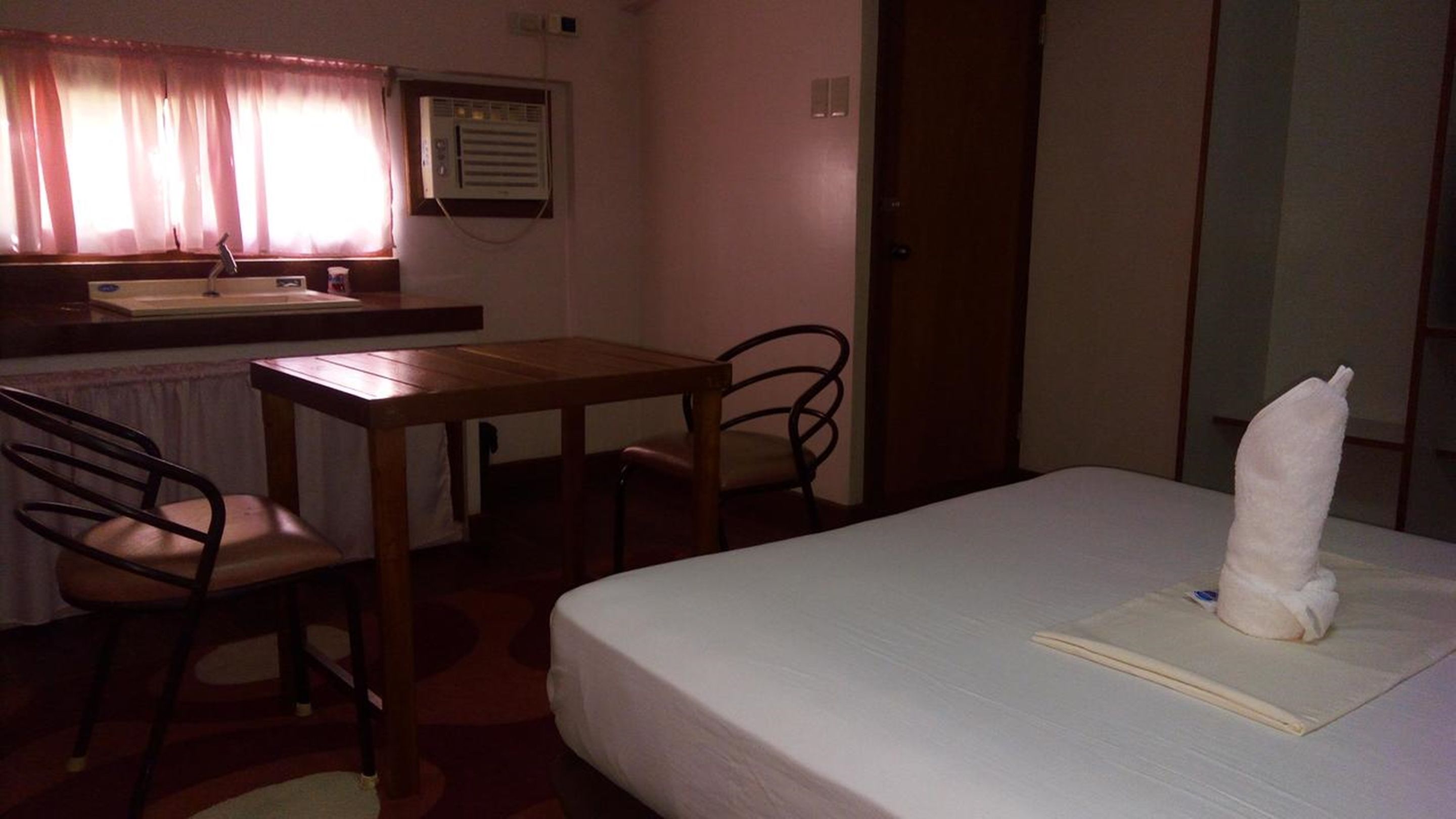 Piccola camera d'albergo con un letto singolo, un tavolo di legno, una sedia, un lavandino e una finestra con tende.