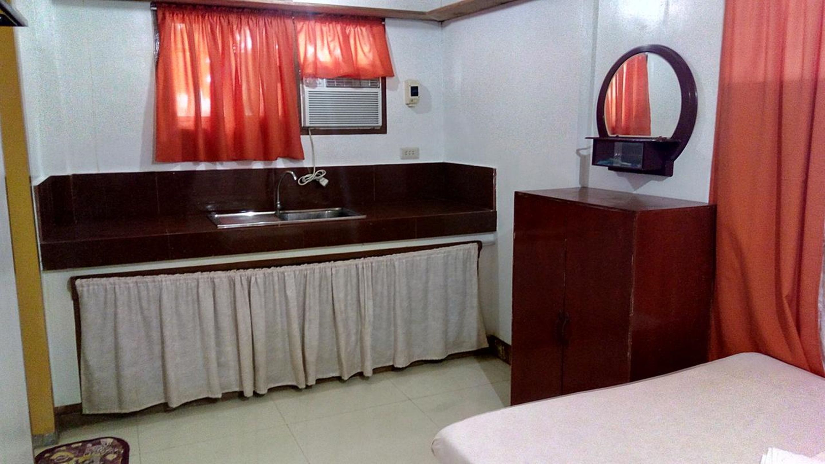 Una habitación pequeña y funcional con un rincón de cocina equipado con pileta y espejo. Se observa un armario y una cama con cubrecama blanco.