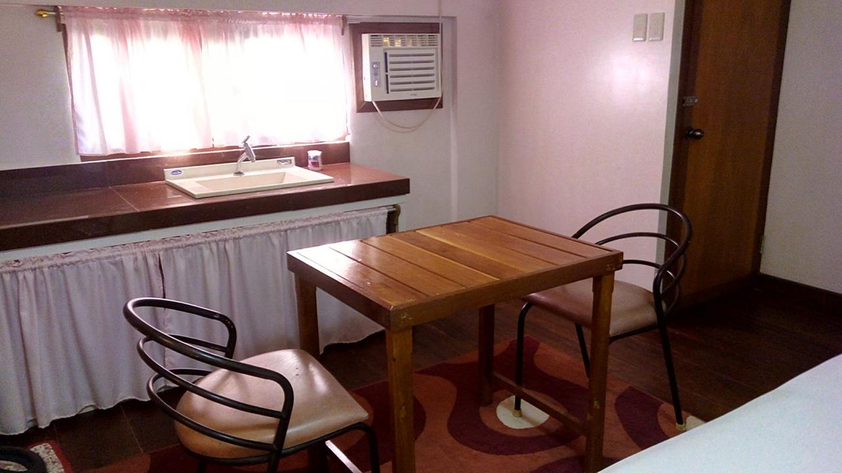 Een kleine kamer met een houten tafel en twee stoelen, een raam met gordijnen, een radiator en een deur.