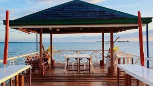 Una terraza de madera con mesas y sillas, un área cubierta con techo verde y vistas al mar.