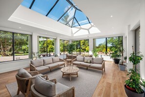Un vaste espace de vie doté d’un haut plafond et de grandes fenêtres, comprenant un canapé d’angle, une table basse ronde et plusieurs plantes en pot.