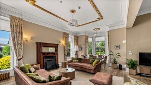 A spacious living room with leather sofas, a fireplace, and large windows offering a view of greenery. - Victorian Mansion, fully refurbished, sleeps 24 guests in 12  ensuite bedrooms (Carnoustie)