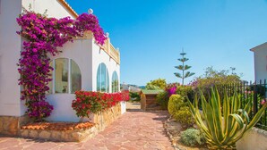 A white building with pink bougainvillea climbing up the wall, a stone pathway, and a cactus plant. - Villa LADERA by Abahana Villas (Calp)
