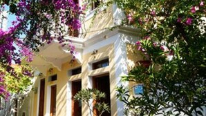 Un edificio de dos pisos con techo rojo, paredes blancas y un balcón. Hay escalones que conducen a la entrada, y el edificio está rodeado de follaje y plantas con flores.