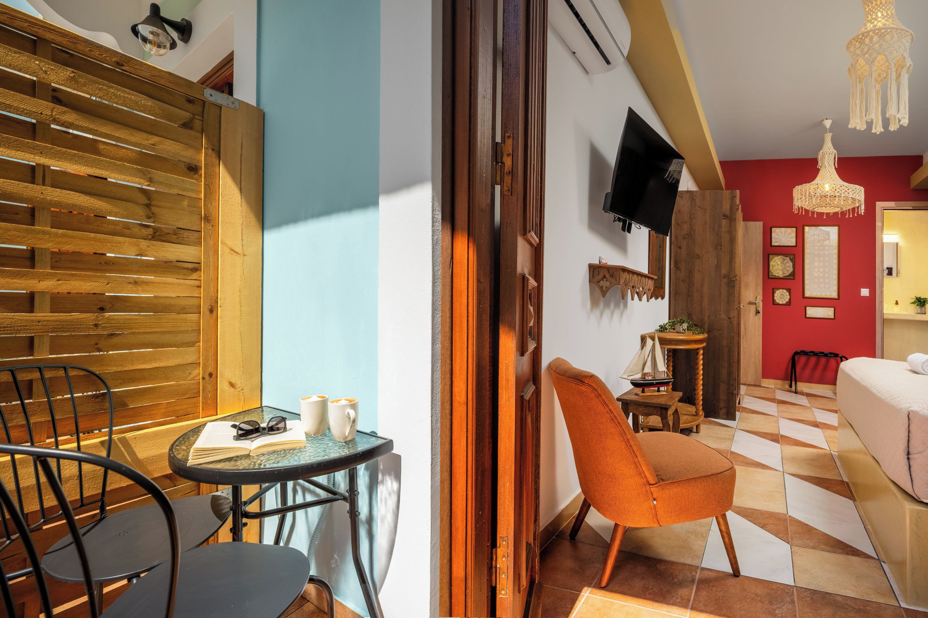 A room with a wooden slatted wall, a small round table with a book and glasses, a single orange chair, a bed with a white cover, and a TV mounted on the wall.