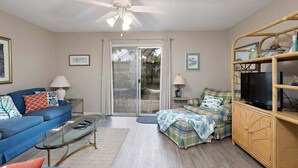 A living room with a blue sofa, a glass coffee table, a wicker cabinet, and a television. - Access to Community Pool & 1 Block to the Beach! (Saint Simons Island)