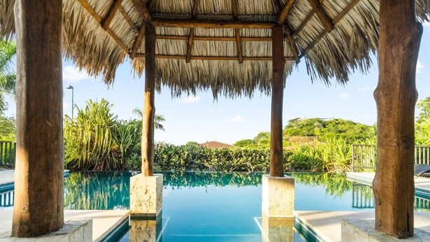 Una piscina infinità amb una estructura de teules de palla, envoltada de vegetació exuberante i palmeres.