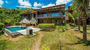 Una casa moderna de dos pisos amb piscina, envoltada de vegetació exuberant i palmeres.
