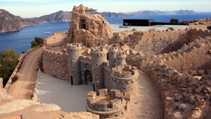 Un castillo de piedra histórico con una entrada fortificada, un camino empedrado y una vista al mar y montañas al fondo.