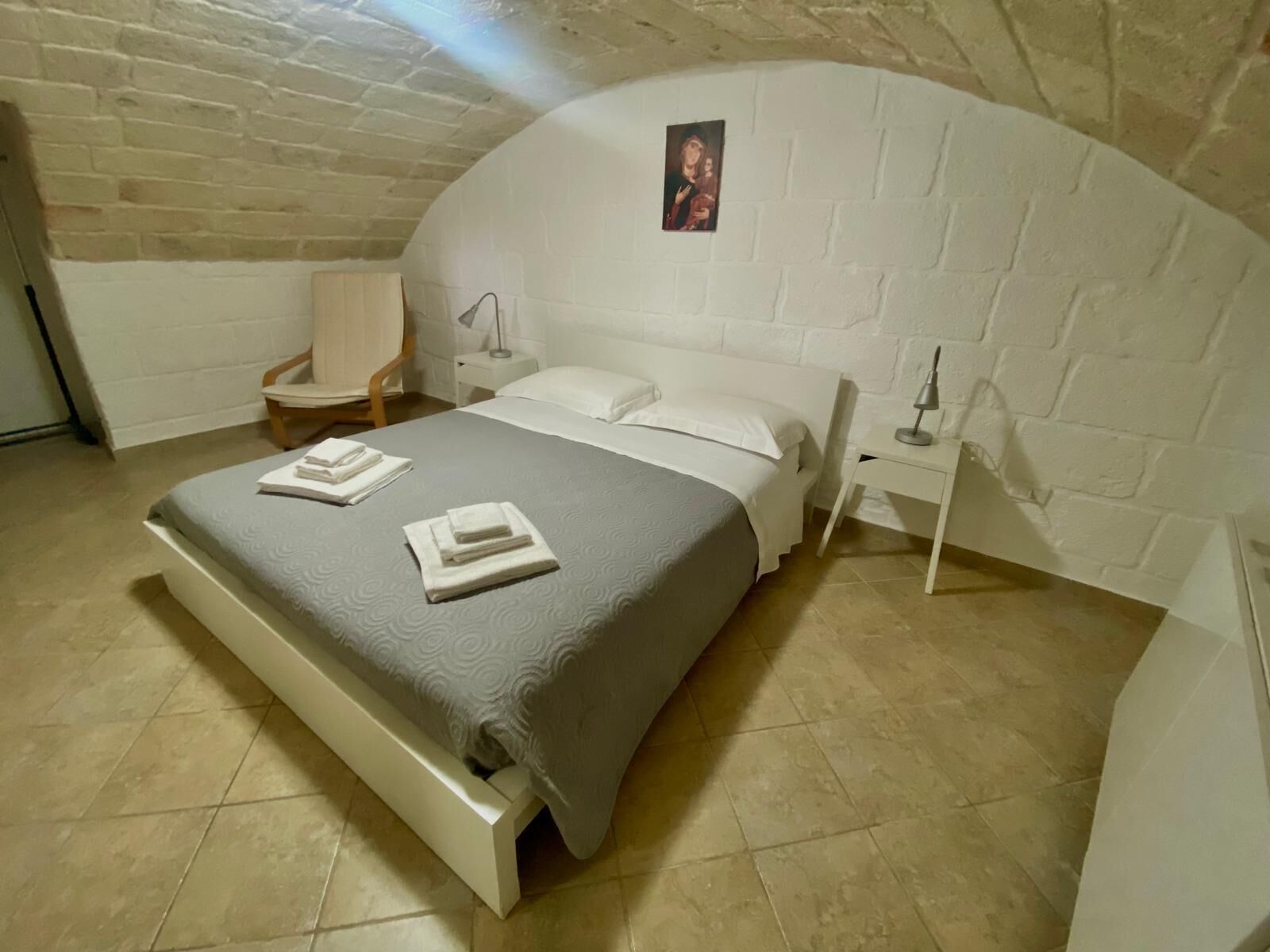 A bedroom with a bed, a chair, a nightstand, and a wall-mounted picture.