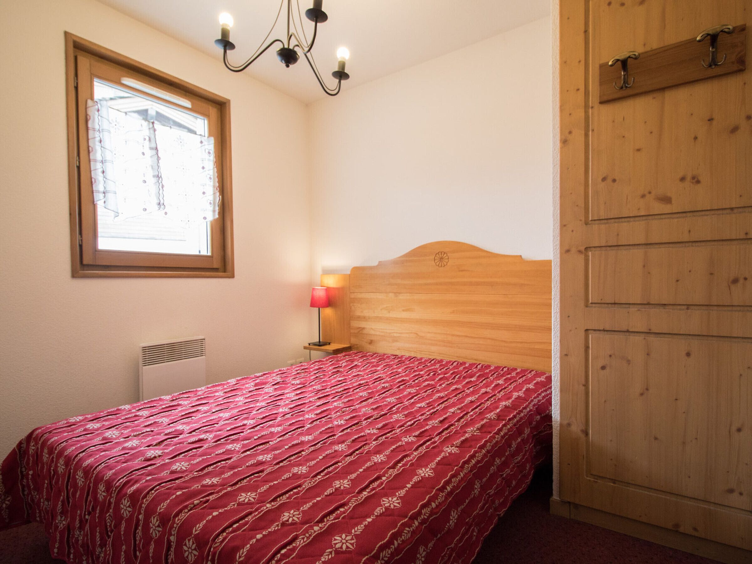Ein Schlafzimmer mit einem Holzkopfteil, einer rot gemusterten Tagesdecke, einer Nachttischlampe und einem Fenster mit Vorhängen.
