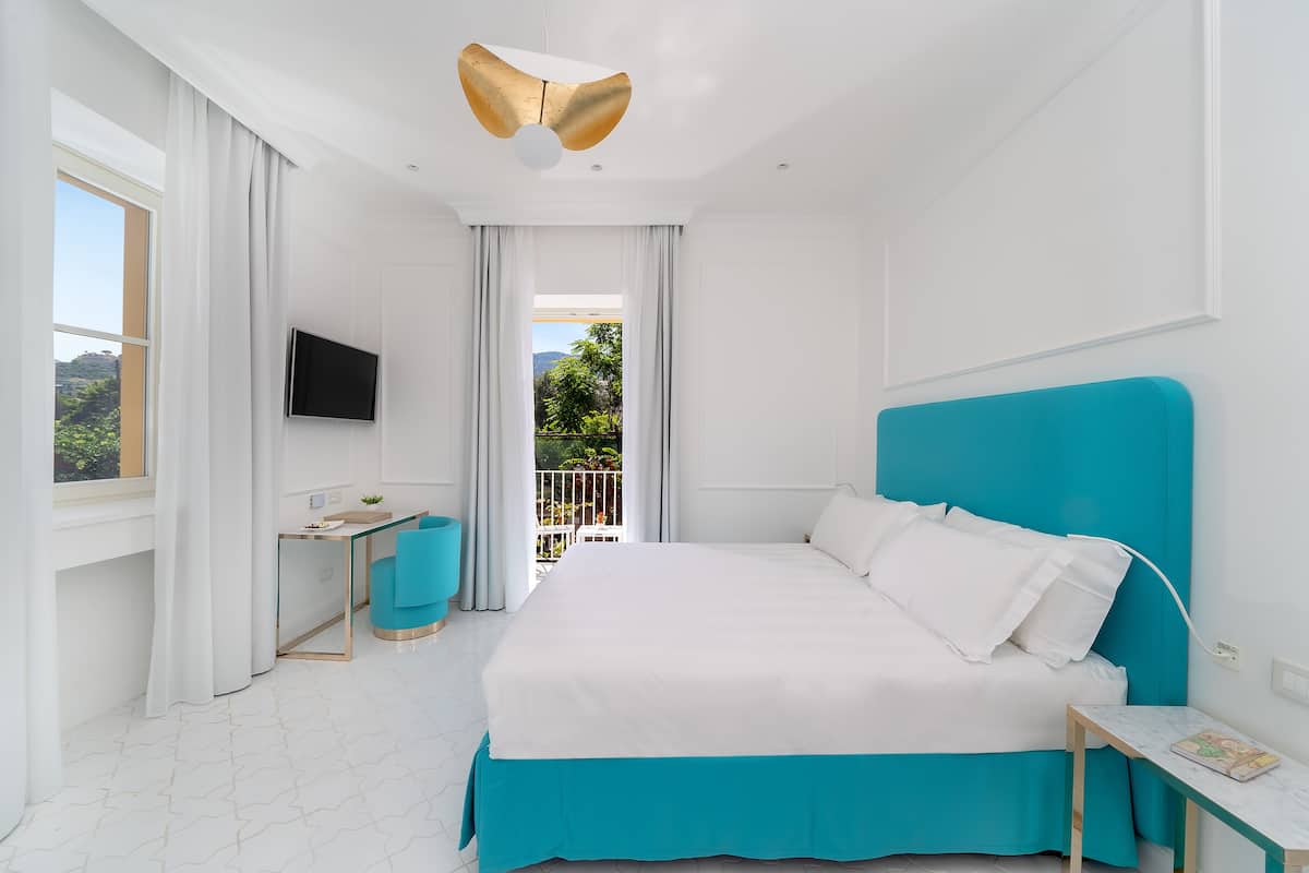 Une chambre d’hôtel moderne dotée d’un grand lit, d’un bureau, d’une chaise, d’une télévision et d’un balcon offrant une vue sur la verdure.