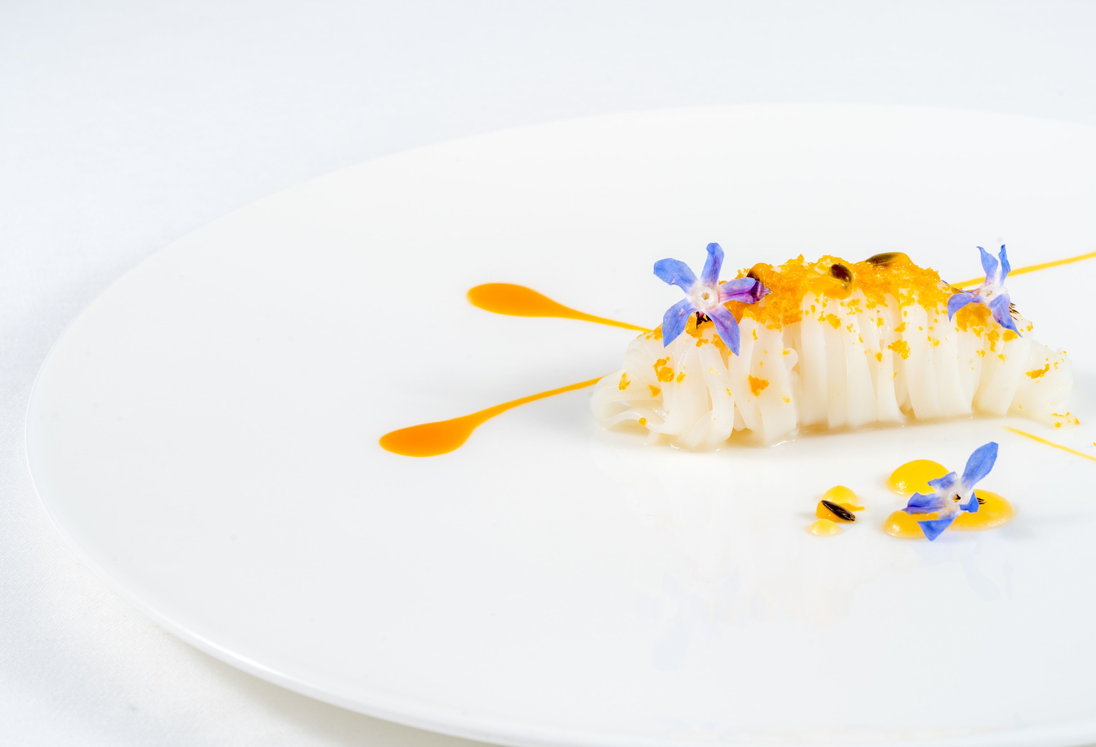 Une assiette blanche sur laquelle est servi un dessert blanc, agrémenté de fleurs bleues et d’une sauce orange.