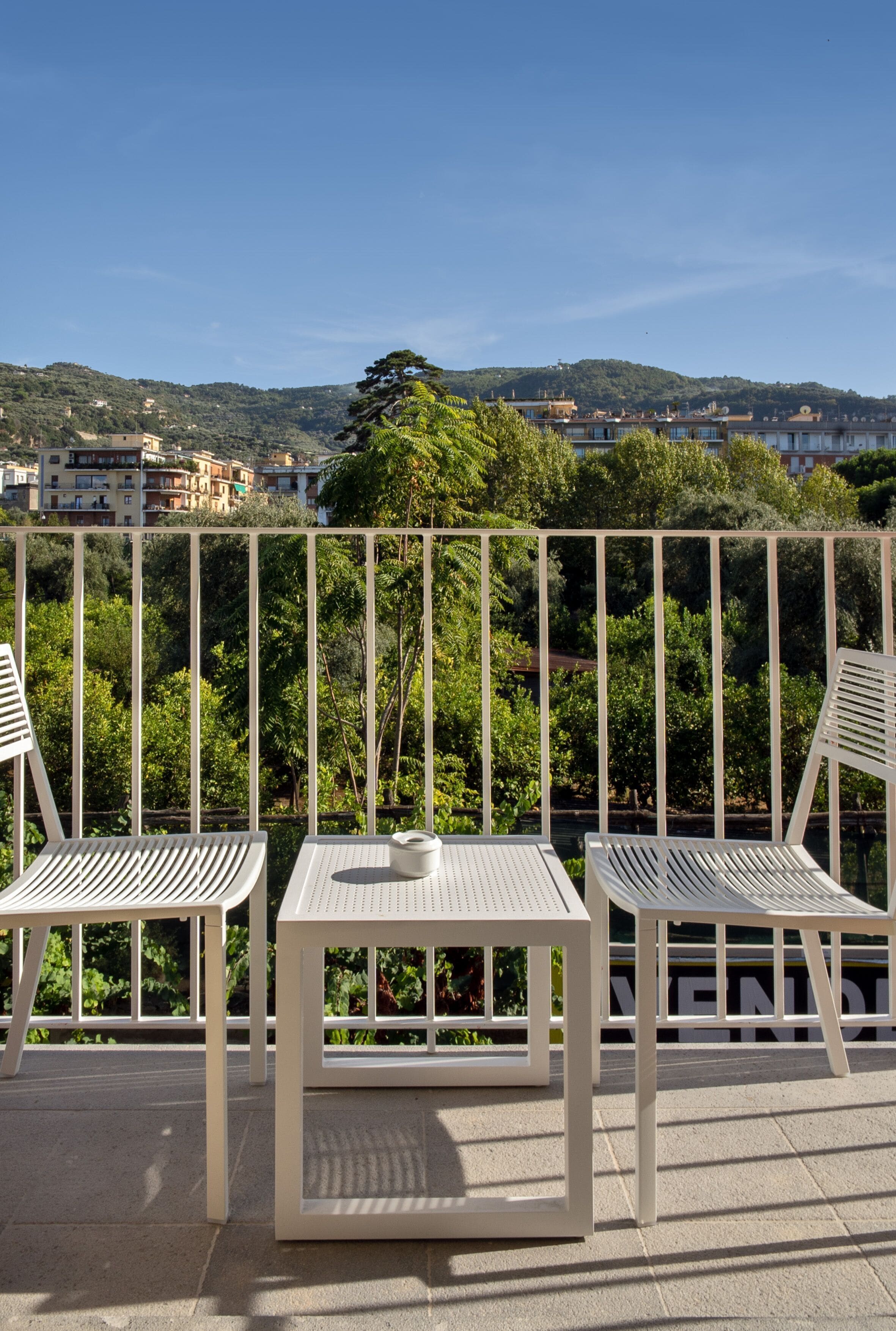 A balcony with two white metal chairs and a small table, overlooking a scenic view of buildings and greenery.