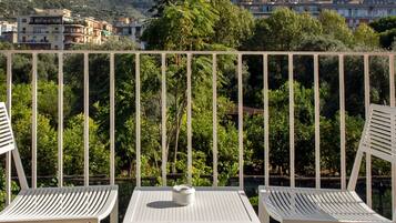 A balcony with two white metal chairs and a small table, overlooking a scenic view of buildings and greenery.