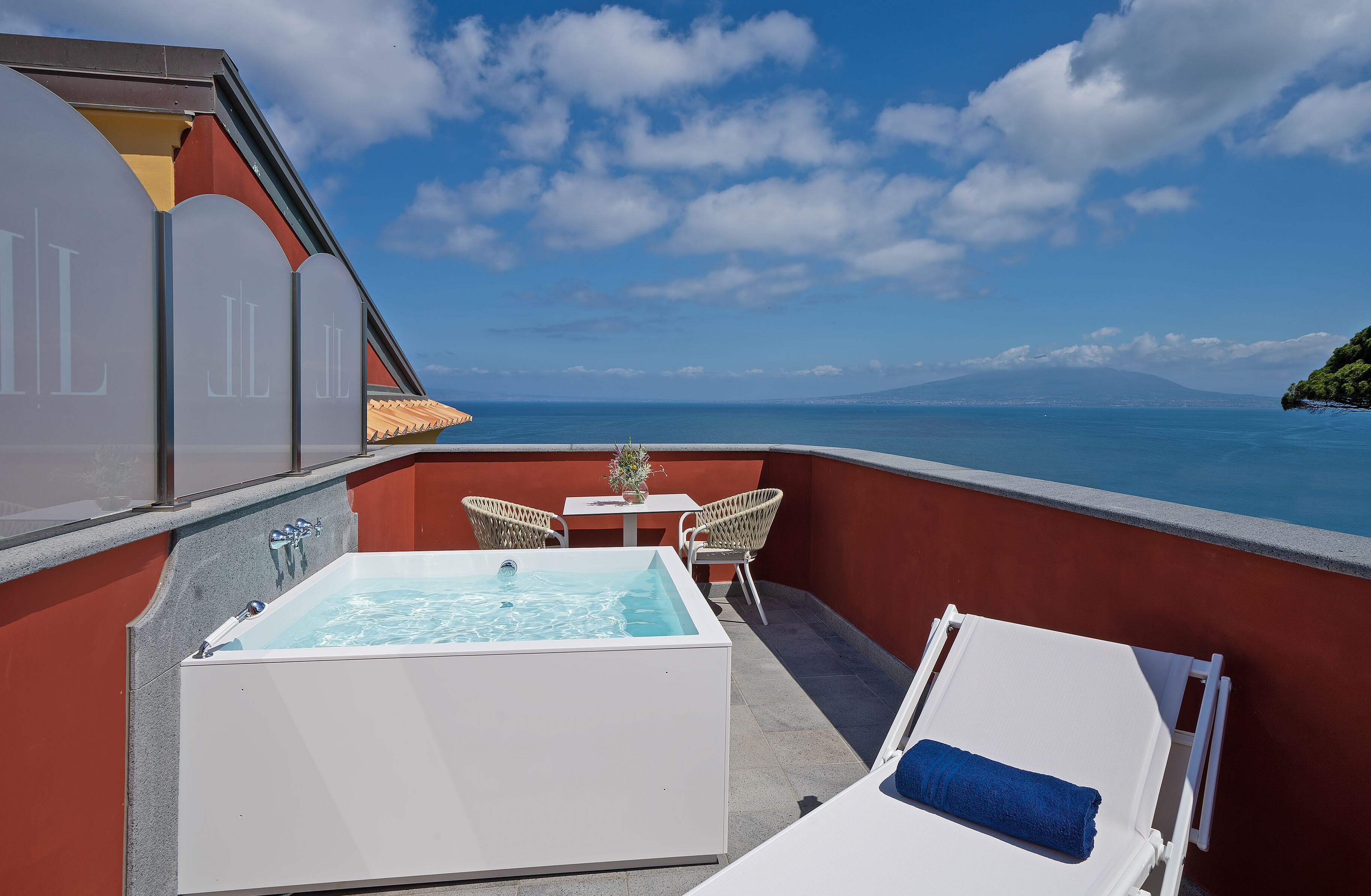 Un balcón con un jacuzzi, dos sillones de mimbre, una mesita con un florero y una tumbona con una toalla.