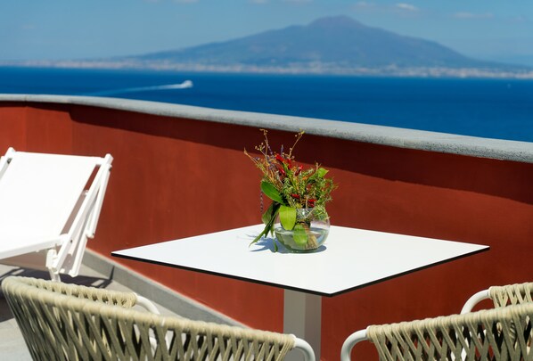 A balcony with a white table and wicker chairs, overlooking a coastal view with a mountain in the distance. - Hotel Lorelei Londres (Sorrento)