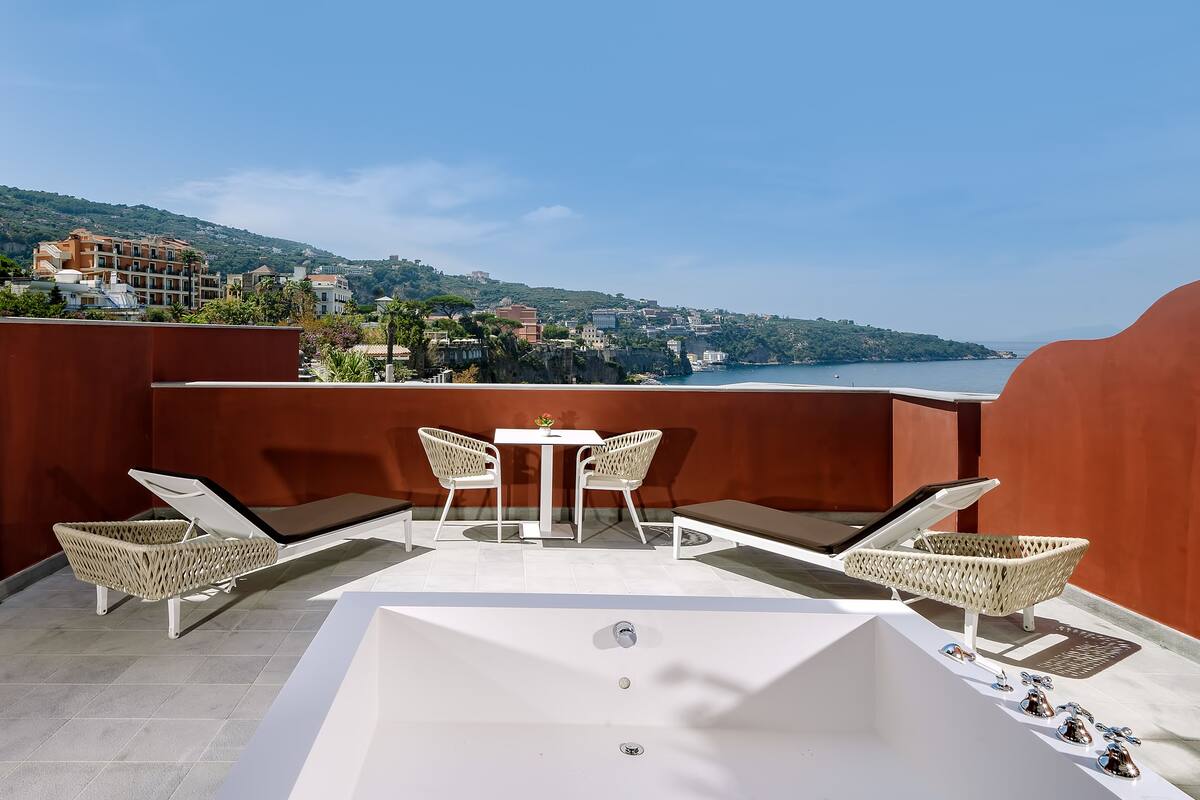 Un balcon avec une baignoire blanche, des fauteuils en osier et une petite table sur laquelle est posé un vase.