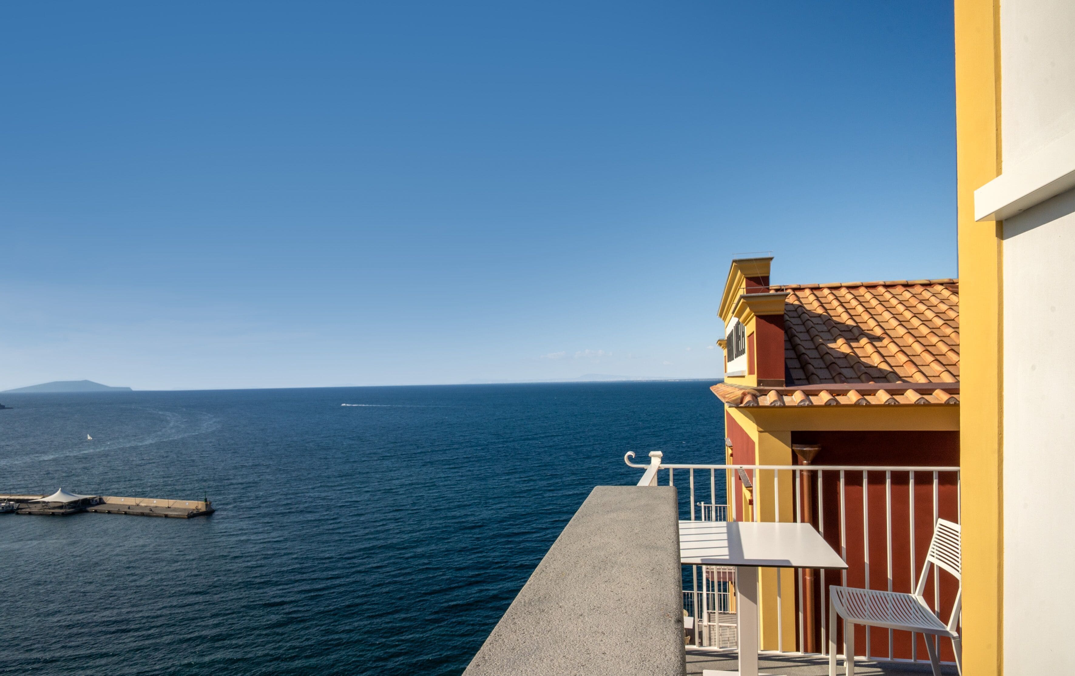A balcony with a view of the sea, a white chair, and a yellow building.