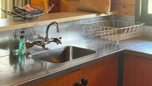 A kitchen with a stainless steel sink, wooden cabinets, a hanging rack, and a framed floral artwork on the wall.