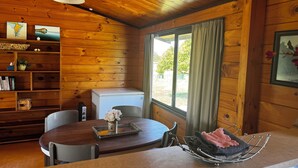 A wooden cabin interior with a dining table, chairs, a small refrigerator, and a window with curtains.