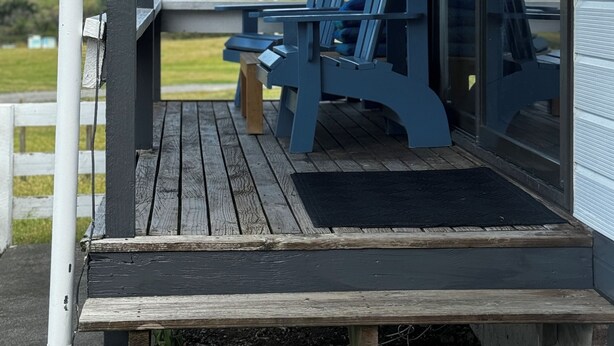 A wooden deck with blue Adirondack chairs and a table, overlooking a grassy landscape with hills.