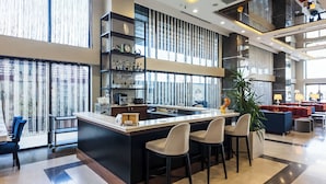 Uma área de bar moderna com balcão, banquetas e máquina de café. Estantes exibem garrafas e copos, e uma grande janela com cortinas.