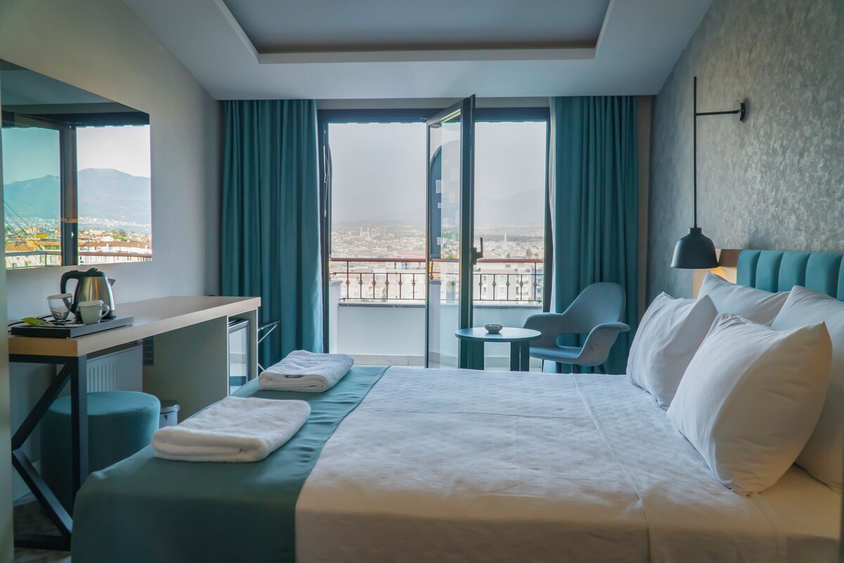 A hotel room with a bed, a desk, a chair, a small table, and a view of the city through large windows.