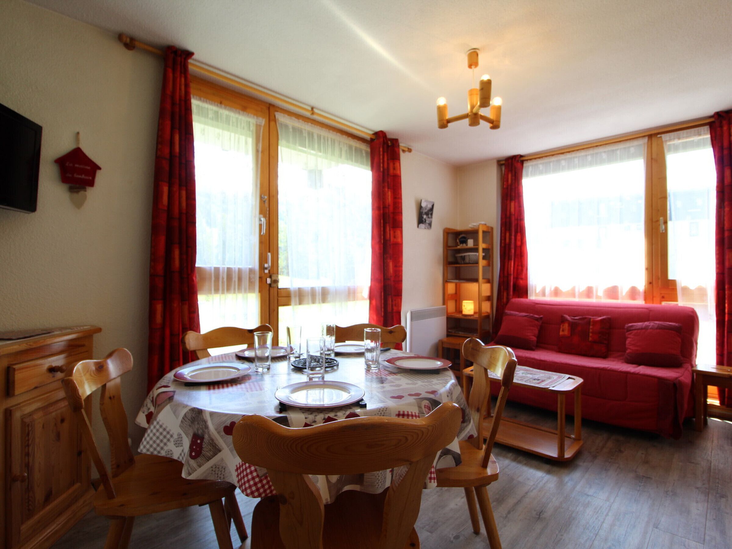 A dining area with a round table set for four, surrounded by wooden chairs. There is a red sofa and a bookshelf in the background.