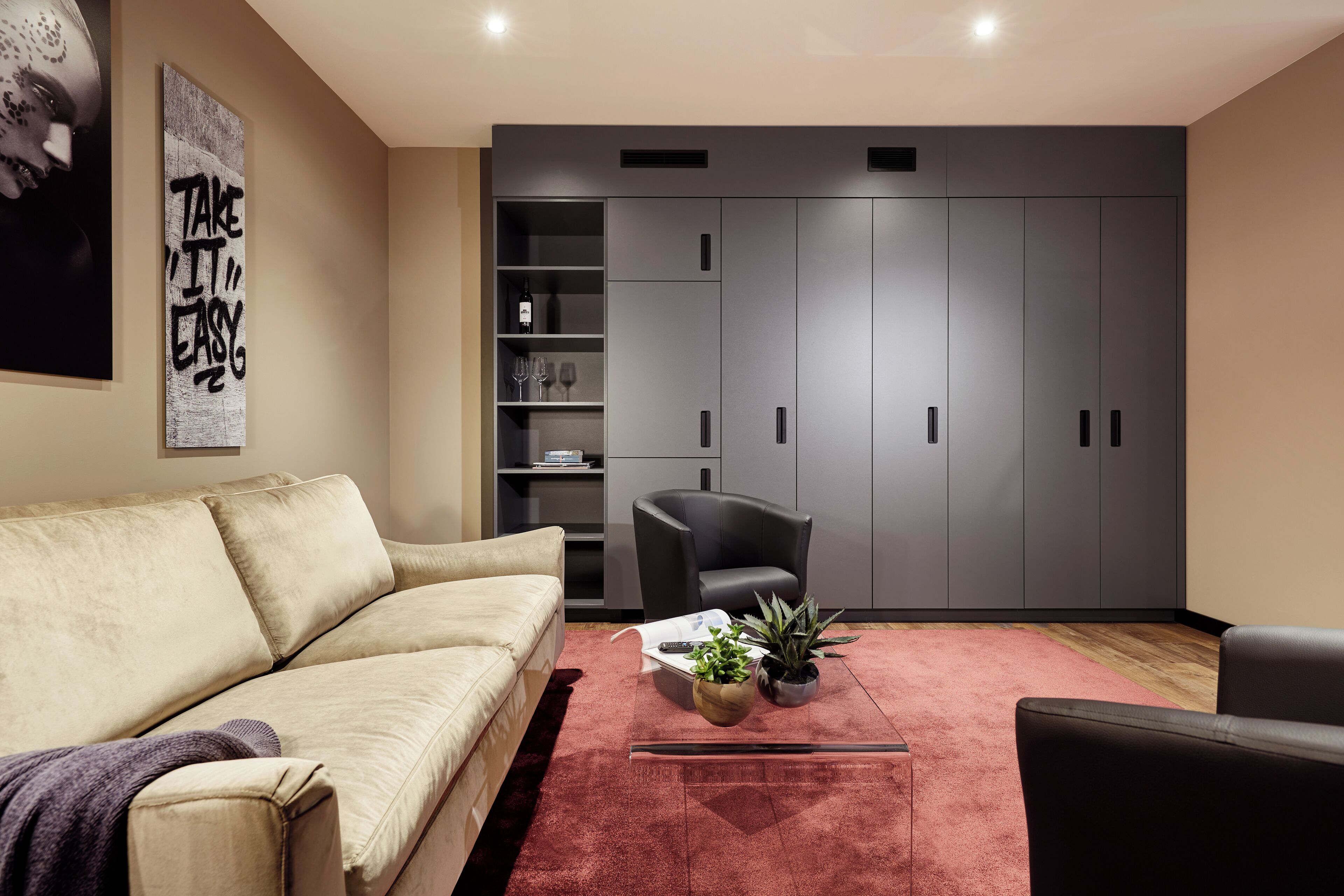 Un salon moderne avec un canapé beige, un fauteuil noir et un tapis rouge. On y trouve une bibliothèque avec des objets décoratifs et une grande armoire grise.