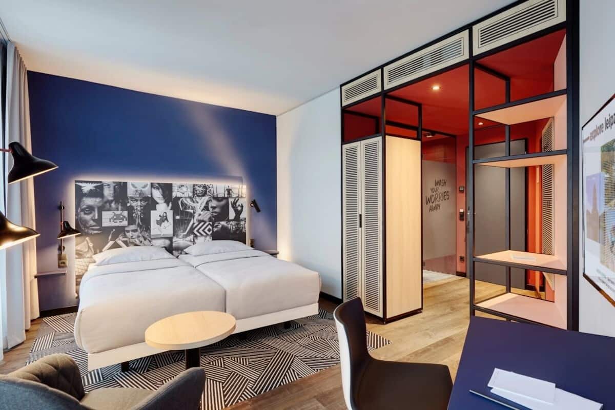 A modern hotel room with a large bed, a desk, and a wardrobe.
