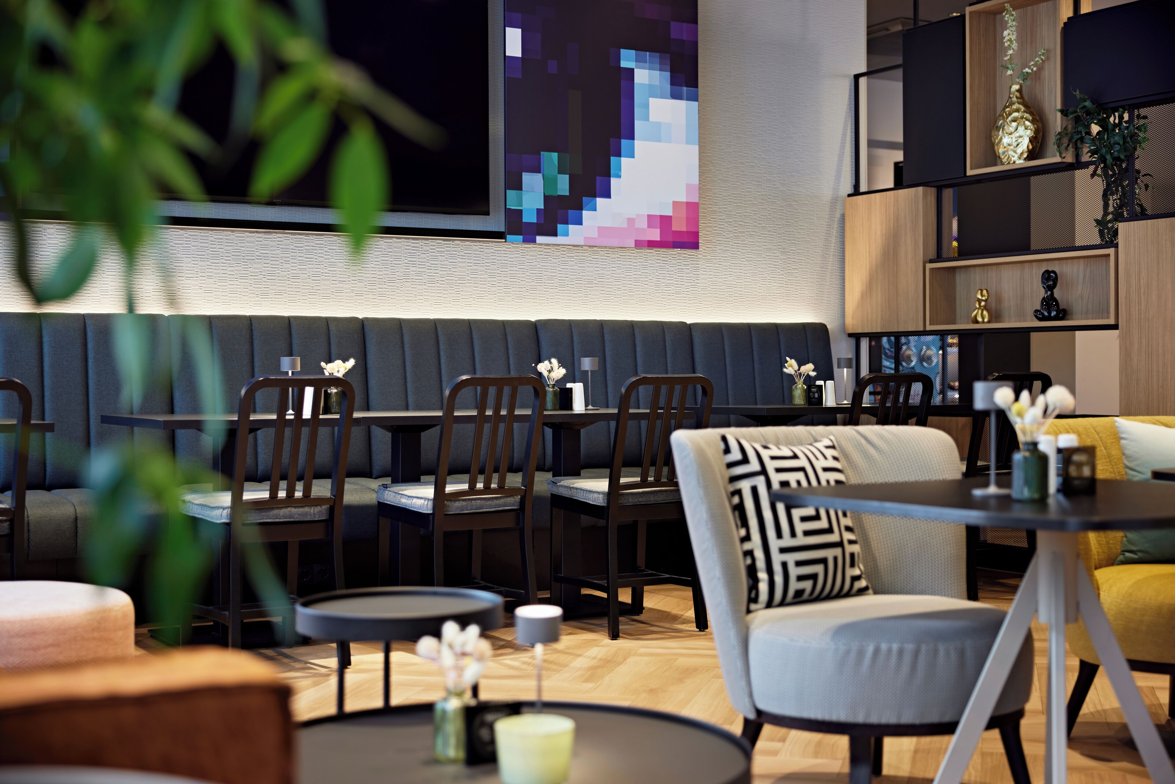 A modern interior with a seating area featuring cushioned benches and chairs, a small round table with a vase of flowers, and a wall-mounted pixel art piece.