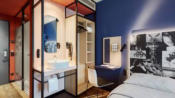 A modern hotel room with a large bed, a desk with a chair, a bathroom with a sink and mirror, and a wall with a large poster.