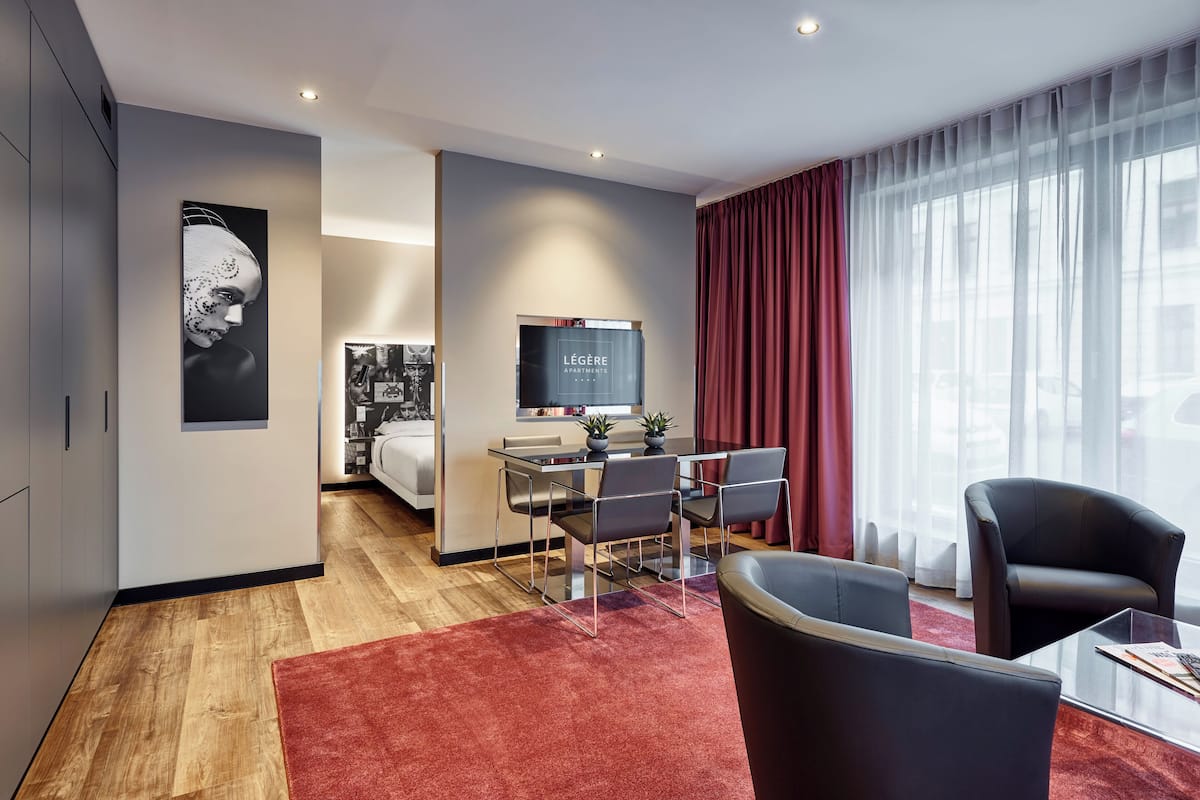 A modern hotel room with a dining area, a large window with sheer curtains, and a flat-screen TV.