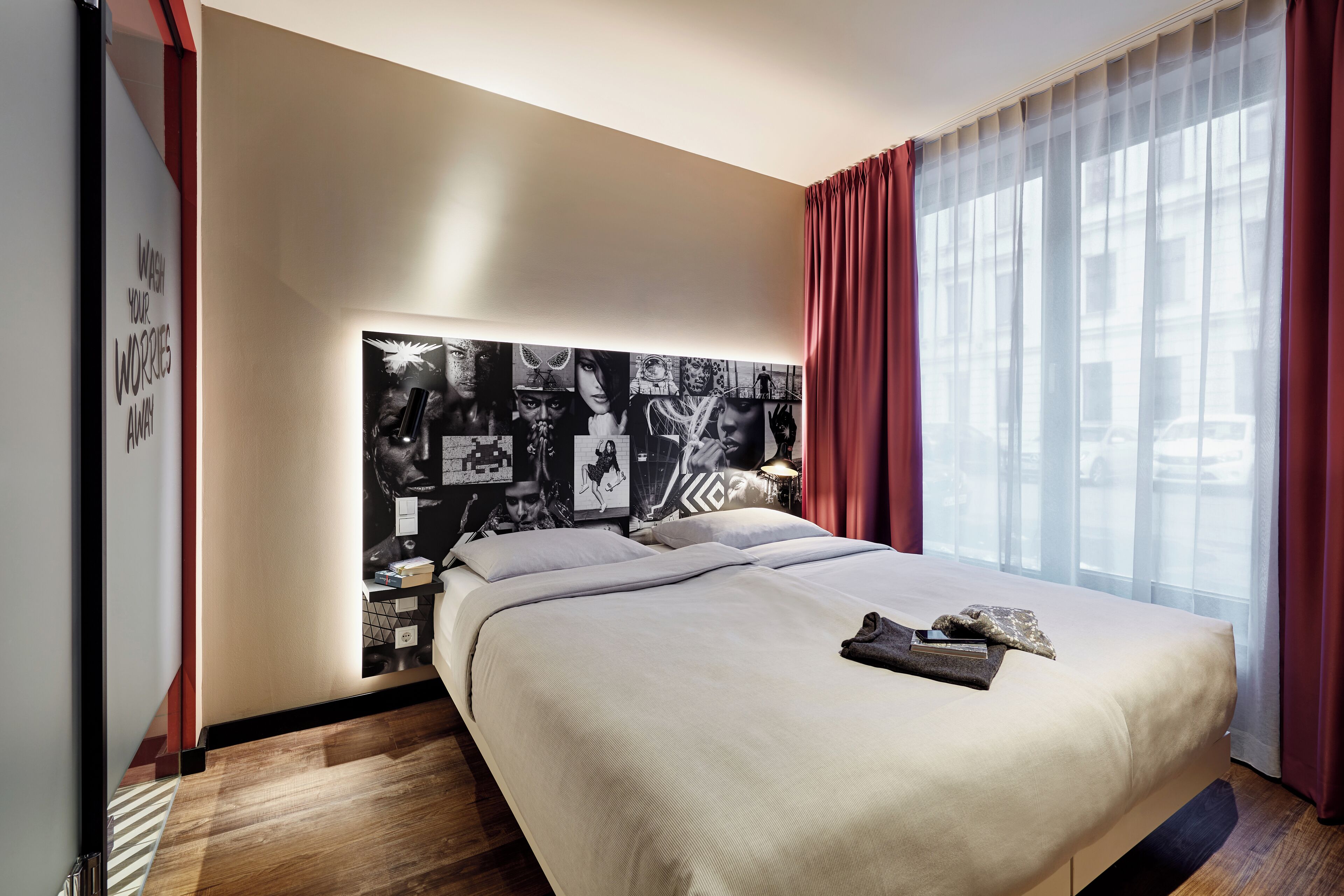 Una habitación de hotel moderna con una cama grande, un cabecero con un collage de fotos en blanco y negro, una ventana con cortinas traslúcidas y un cartel motivacional en la pared.