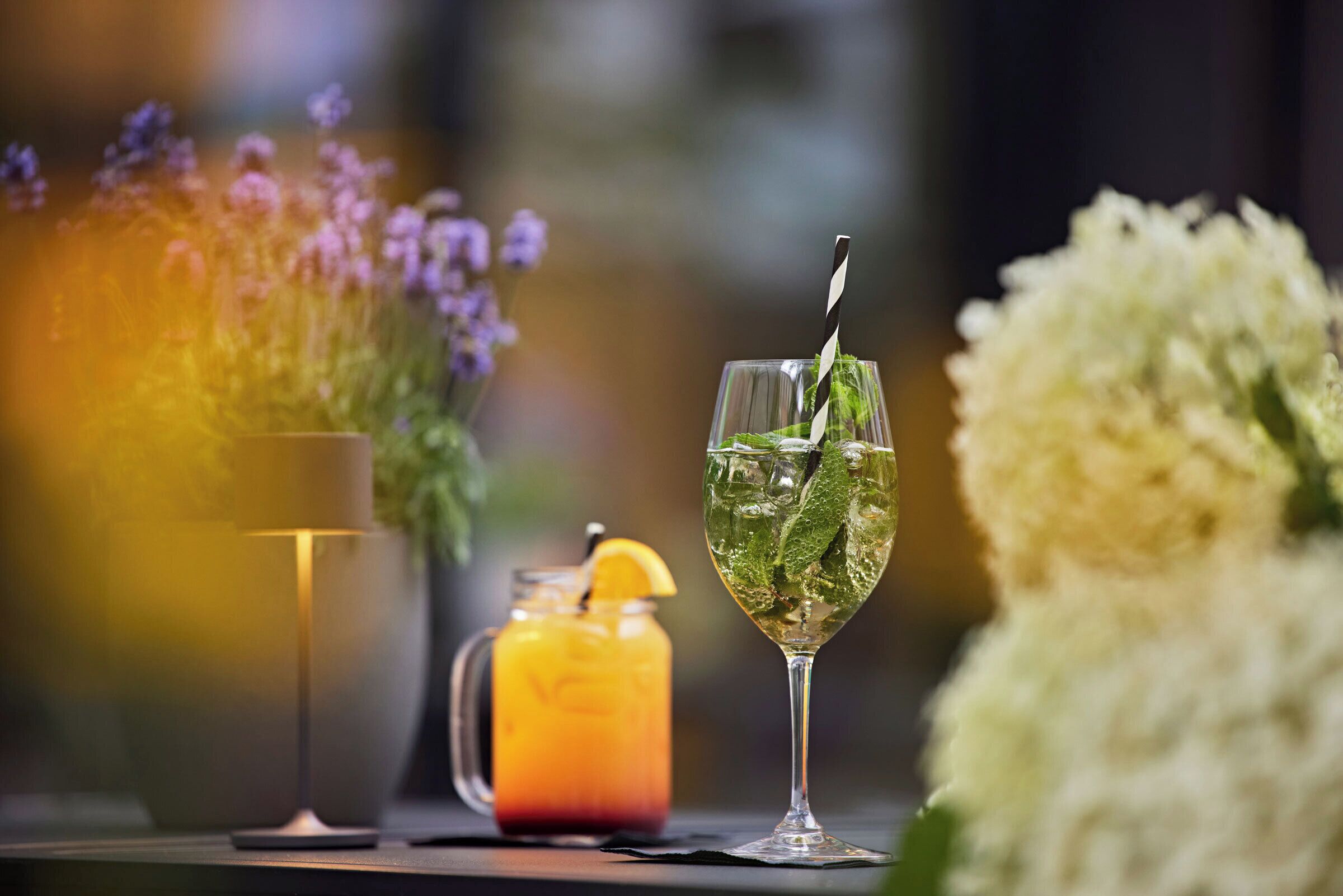 A table with a glass of iced drink garnished with mint and an orange slice, a pitcher of iced tea, and a vase of flowers.