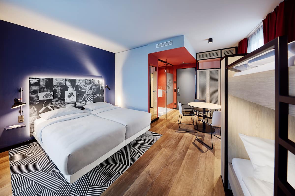 A modern hotel room with a large bed, a desk with a chair, and a bunk bed.
