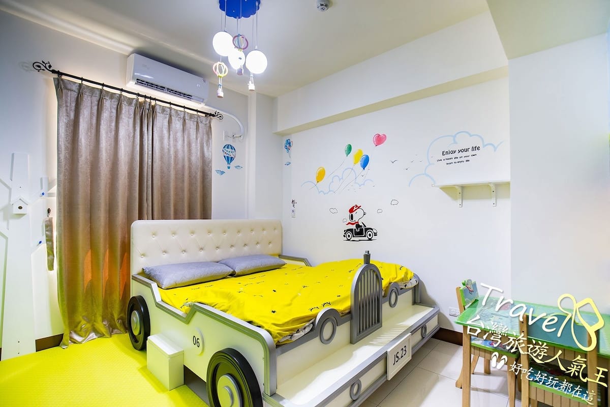 A child's bedroom with a bed designed like a car, a window with curtains, and wall decorations including balloons and a hot air balloon.