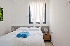 A neatly made bed with white linens and two rolled blue towels, a bedside lamp, and a window with sheer curtains.