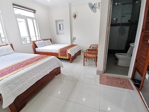 A hotel room with two beds, a sitting area with a chair, a small table, a fan, and a view of the bathroom.