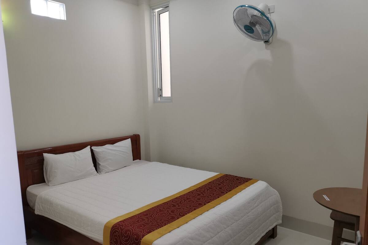 Un lit simple avec une tête de lit en bois, un couvre-lit blanc et un chemin de table décoratif rouge et jaune. On aperçoit un ventilateur fixé au mur, et une petite table ronde se trouve à droite du lit.