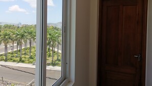 A room with a large window, a wooden door with a number plate, and a view of palm trees and a street.