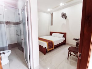 A hotel room with a glass-enclosed shower, a bed with a white and red bedspread, a small table with a chair, and a wall-mounted fan.
