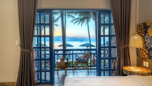 Ett hotellrum med ett stort fönster med utsikt över en strand, en säng, en stol och ett litet bord med en skål med frukt.