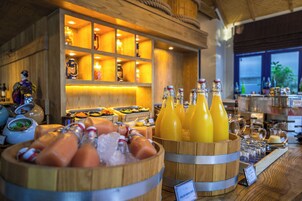 Zona de bufé con diversos alimentos y bebidas, incluyendo un barril de madera con botellas de jugo de naranja y un letrero que indica 'Jugo de Guayaba Helado'.